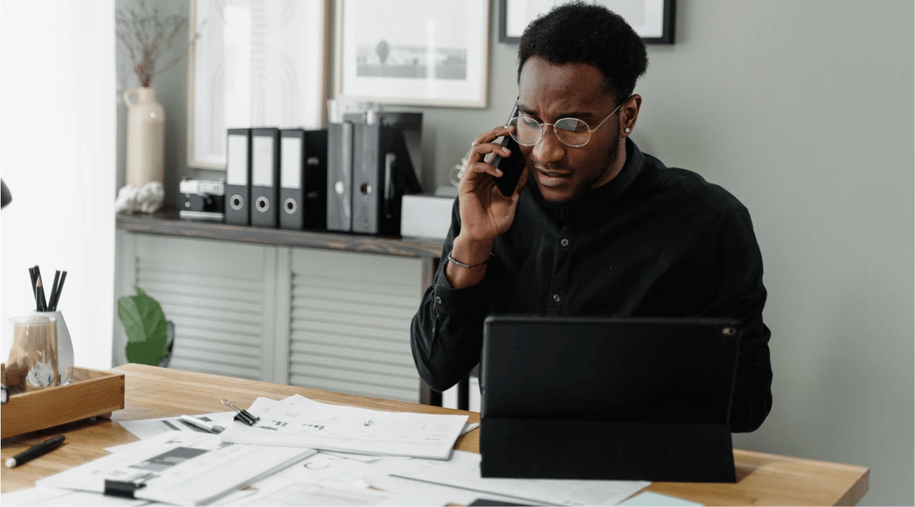 Picture of a businessman in an office. The man is dressed in black, wearing stylish glasses, and talking over the phone while sitting at a desk with a laptop and papers on it.