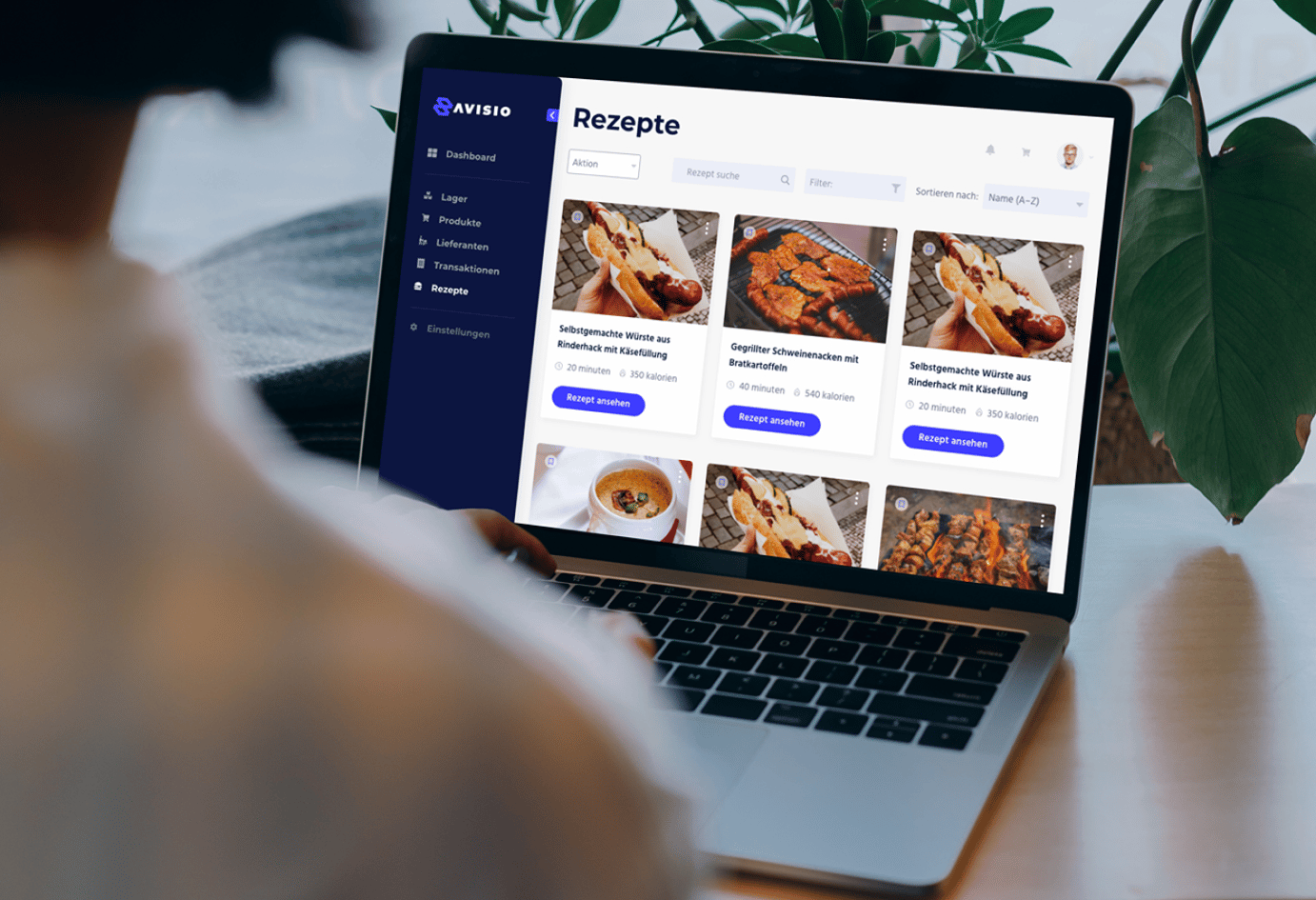 Picture of a laptop on a desk with a plant in the background and a person's back in the foreground. On the laptop screen there is a view from the Avisio hotel inventory management application built by Monterail.