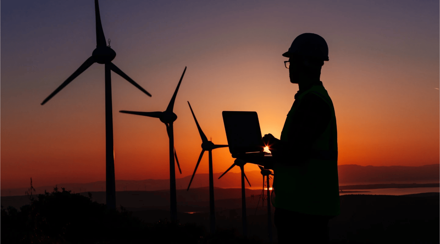Picture of a man in a hard hat standing in front of power-generating windmills at sundown while holding a tablet.